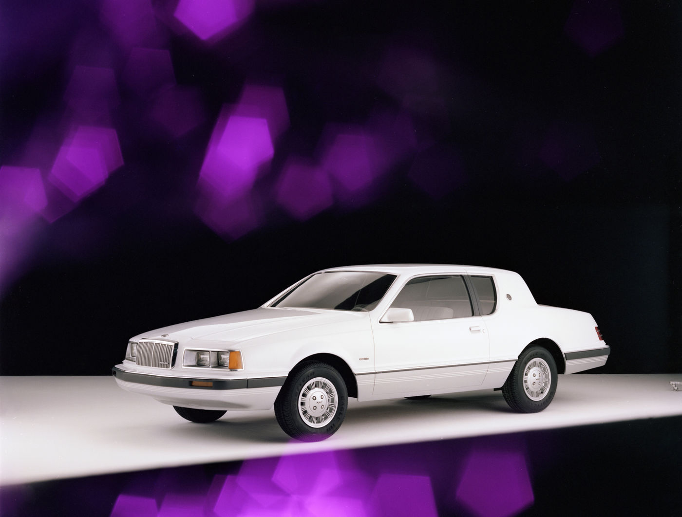 White 1984 Mercury Cougar XR-7 coupe on a white stage with purple blurred lights behind, front three-quarter view