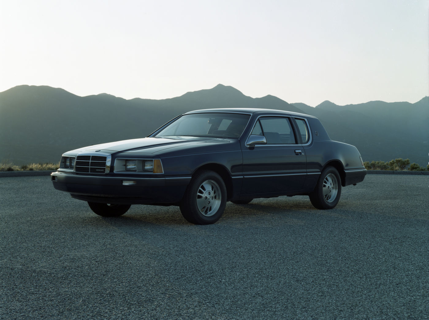Black 1985 Mercury Cougar coupe on a paved overlook with mountains behind, front three-quarter view