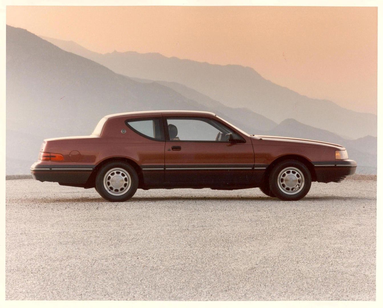 Dark red 1986 Mercury Cougar coupe in full side profile on a gravel lot with hazy mountains at sunset
