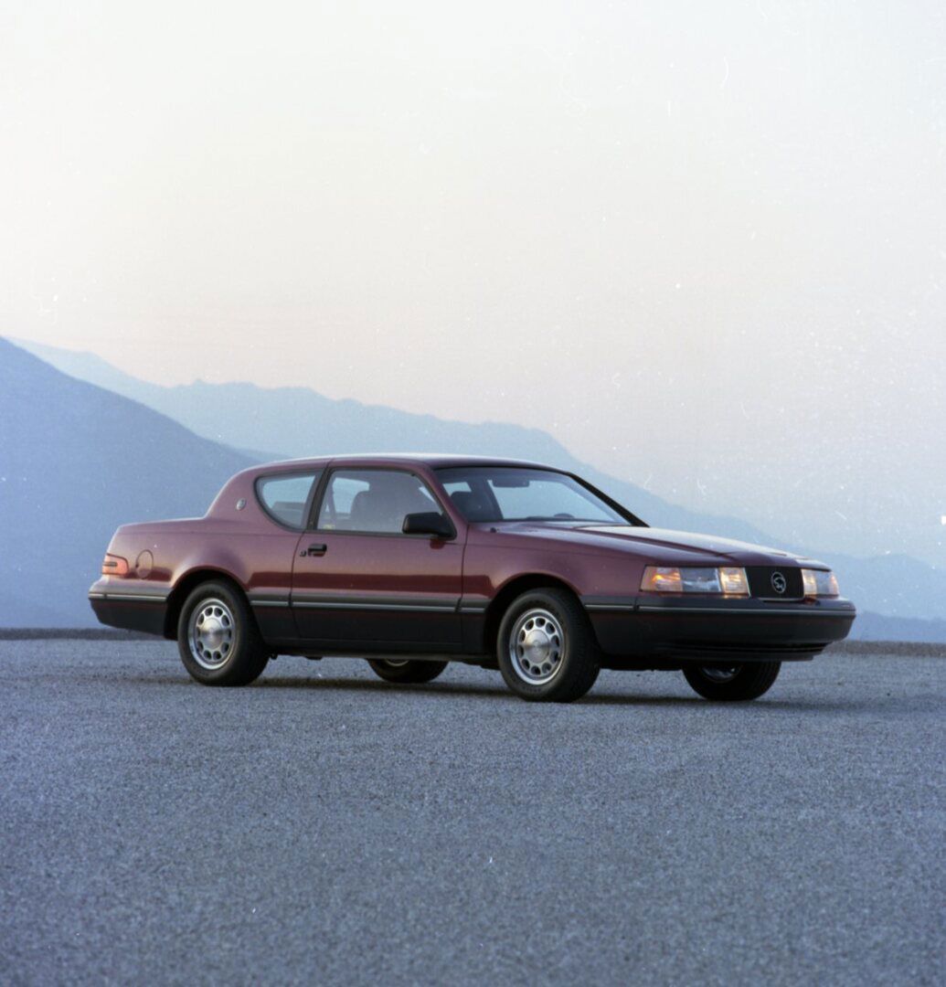 Dark red 1987 Mercury Cougar coupe on a paved lot with misty mountains, front three-quarter view