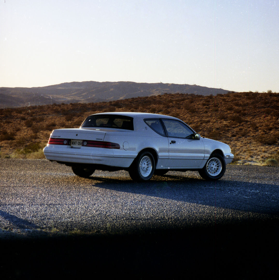 White 1988 Mercury Cougar XR-7 coupe seen from the rear three-quarter on a gravel road through brown hills