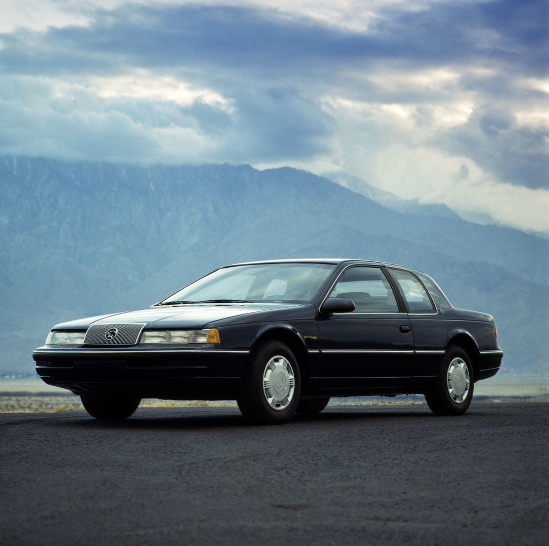 Black 1989 Mercury Cougar XR-7 coupe on a paved lot below cloud-capped mountains, front three-quarter view