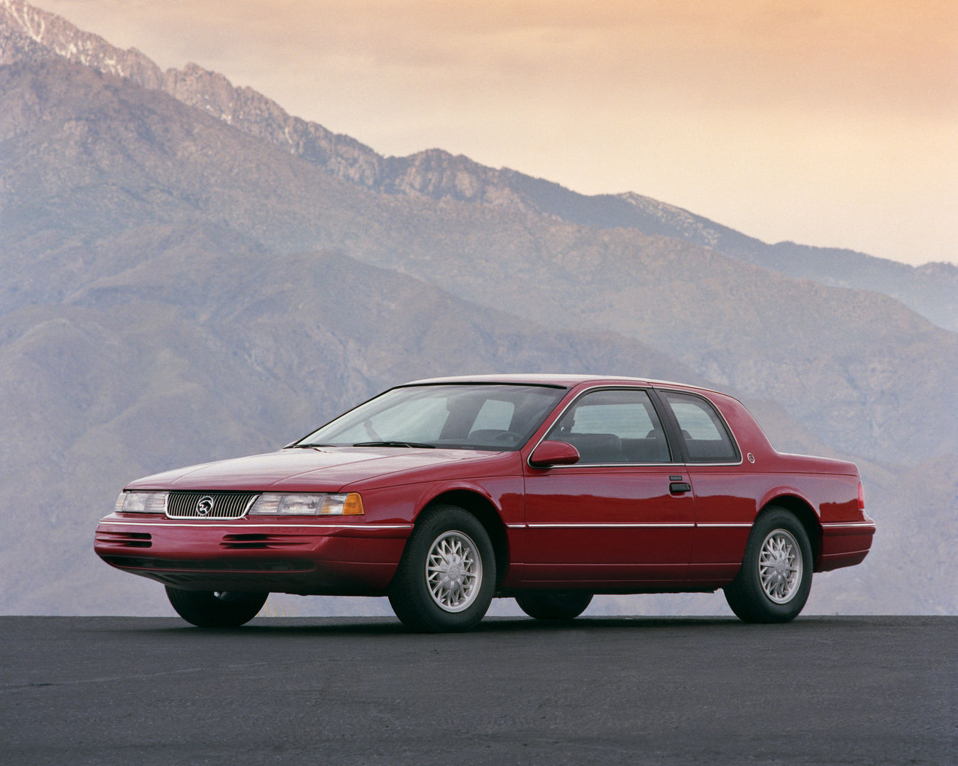 Red 1993 Mercury Cougar coupe on a dark apron with mountains behind, front three-quarter view