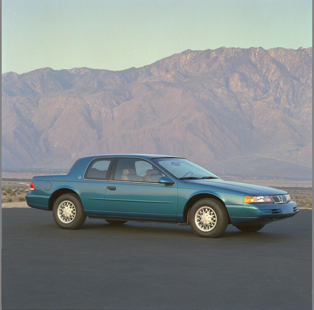Teal 1994 Mercury Cougar coupe in side and front three-quarter view on a road below a mountain range