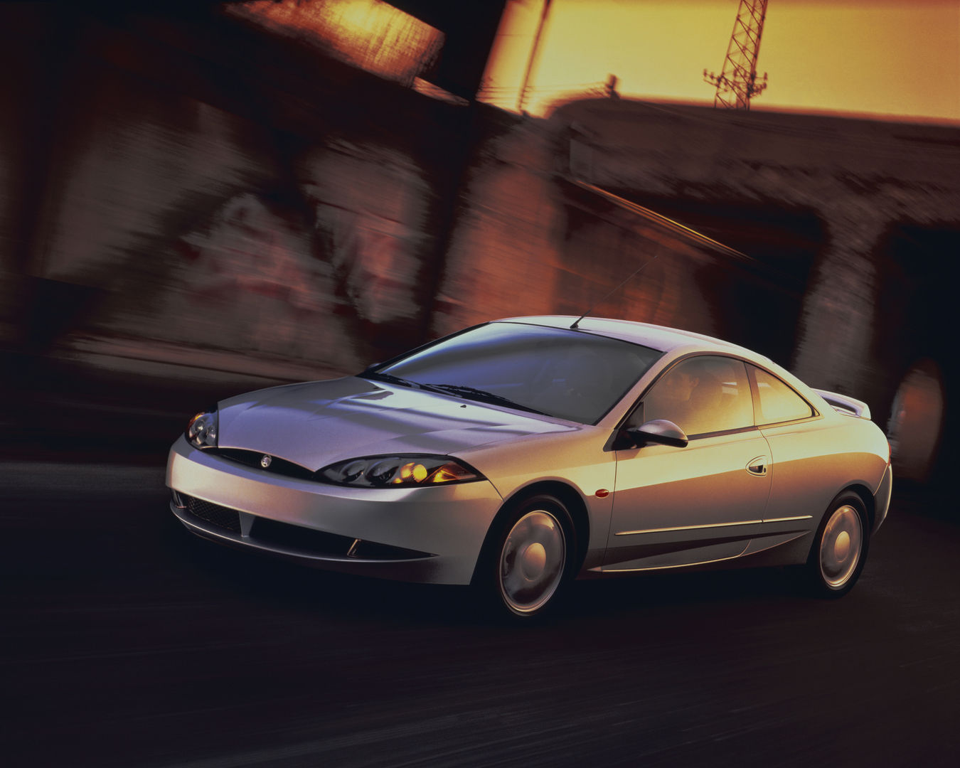 Silver 1999 Mercury Cougar hatchback coupe driving past a blurred building at sunset, front three-quarter view