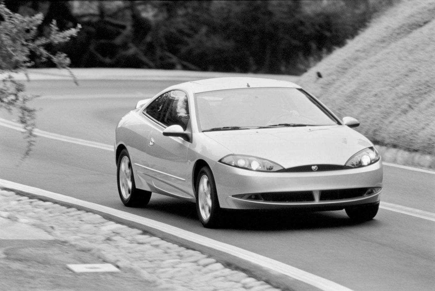 Black-and-white photo of a light-colored 2000 Mercury Cougar hatchback coupe on a curving road, front three-quarter view