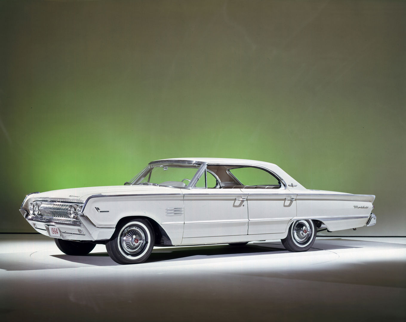 White 1964 Mercury Marauder four-door hardtop with the slanted fastback roof, front three-quarter studio view on a green-lit backdrop