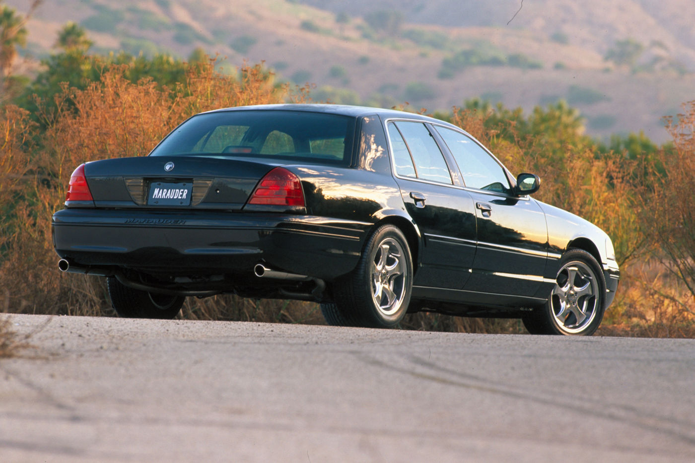 Black 2003 Mercury Marauder sedan seen from the rear three-quarter on a hillside road at sunset, with the MARAUDER display plate and dual exhausts