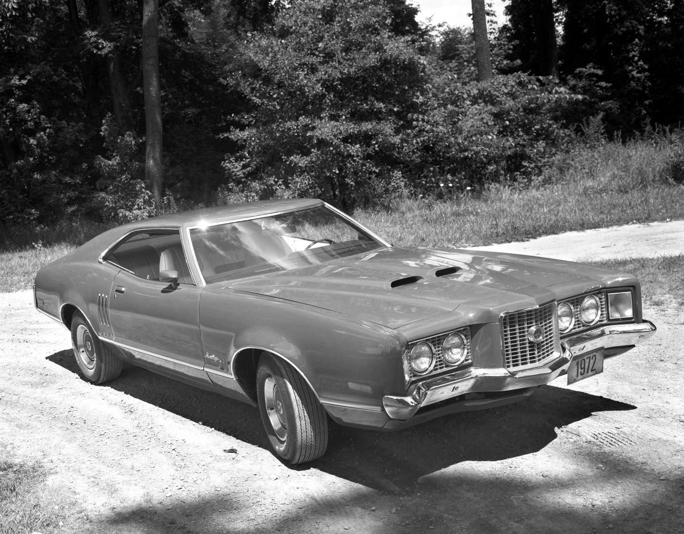 Black-and-white photo of a 1972 Mercury Montego GT fastback on a gravel road, high front three-quarter view, showing the twin hood scoops, fender vents and the slim 1972 bumper