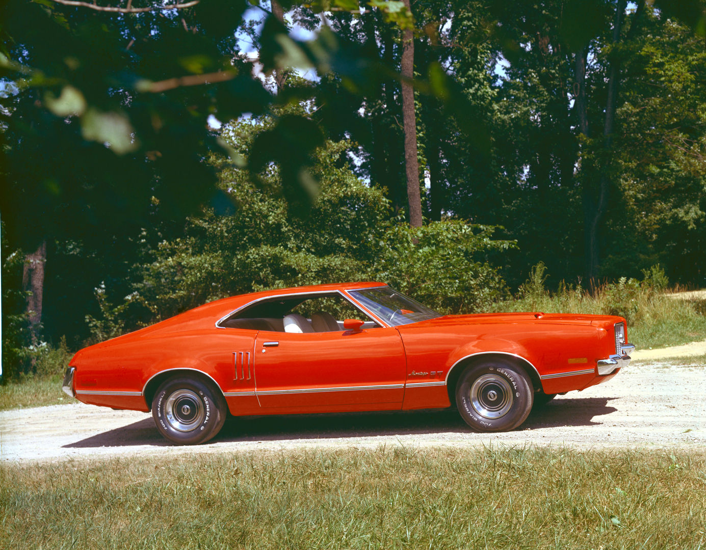Red-orange 1972 Mercury Montego GT fastback hardtop in side profile on a gravel road in front of dense woods