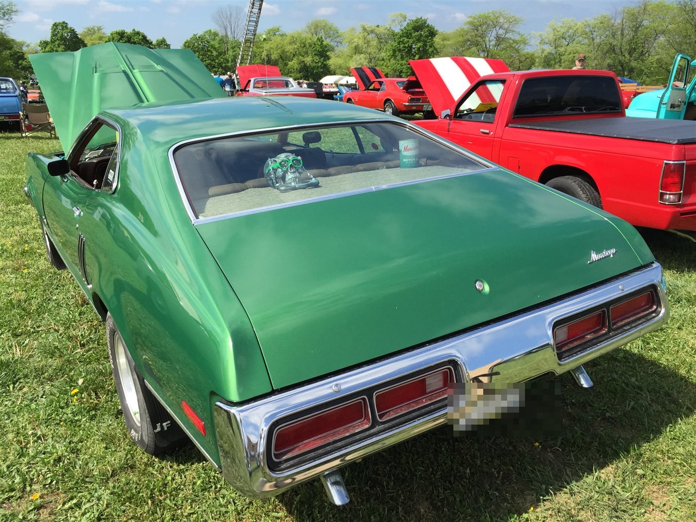 Green 1973 Mercury Montego GT fastback, rear three-quarter view with hood and trunk open, parked on grass among other show cars at a 2015 Antique Automobile Club of America meet; a novelty skull ornament sits on the rear parcel shelf