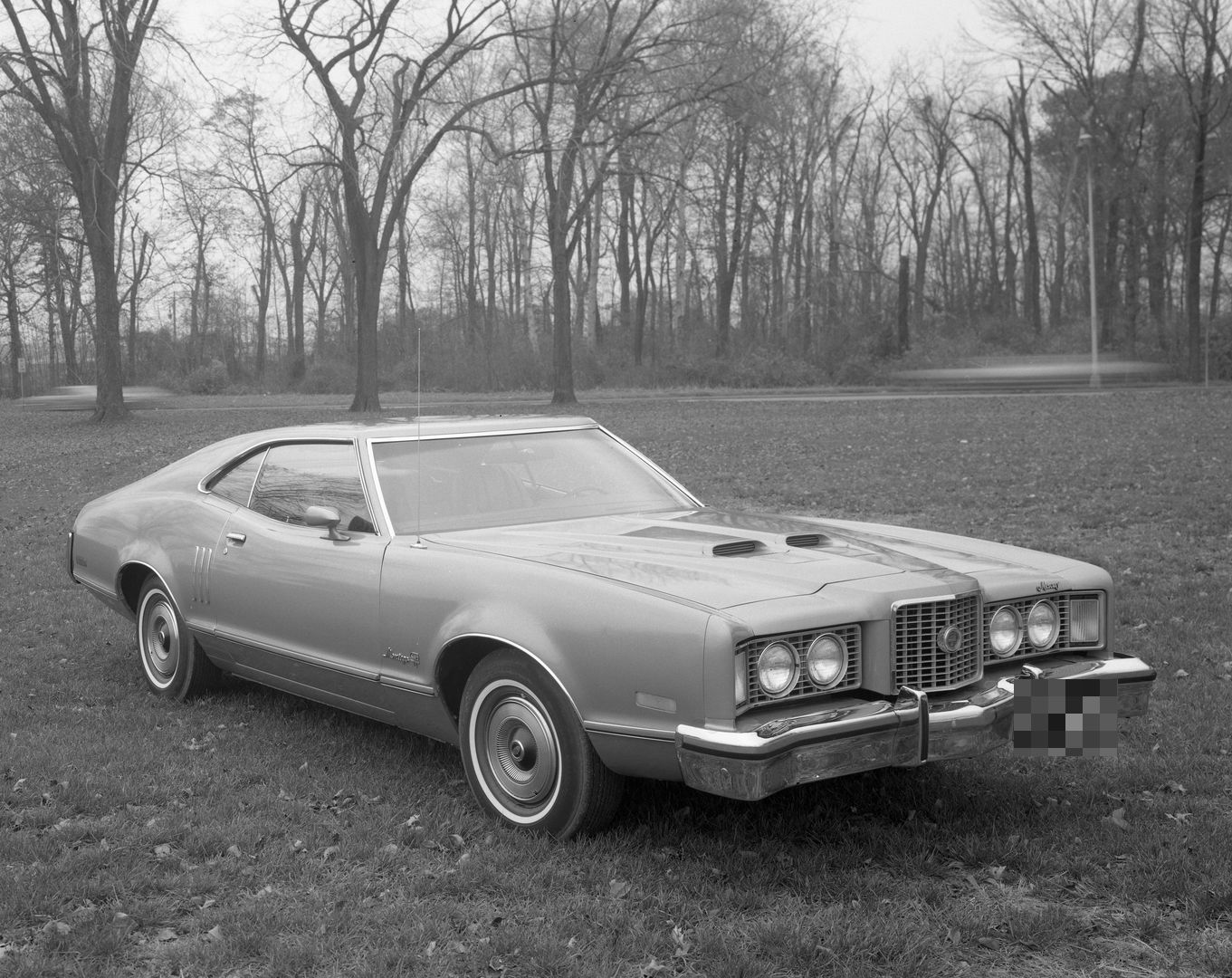 Black-and-white photo of a 1973 Mercury Montego GT fastback on a lawn with bare trees, front three-quarter view, with twin hood scoops and a heavier front bumper with rubber-faced guards