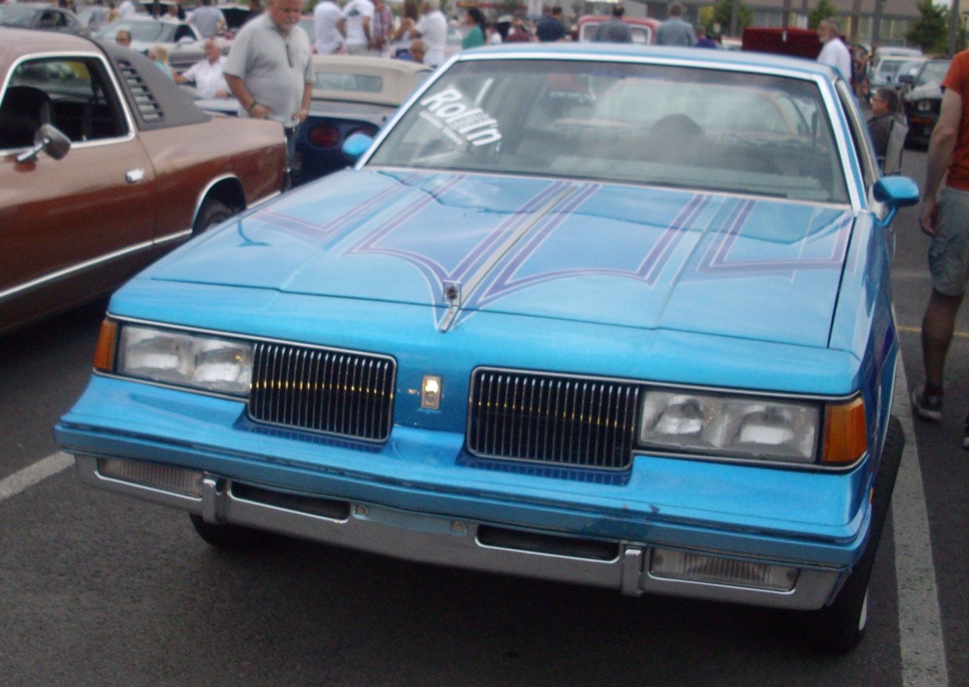 Front view of a blue 1988 Oldsmobile Cutlass Supreme Classic coupe with custom pinstriped hood graphics at a car show