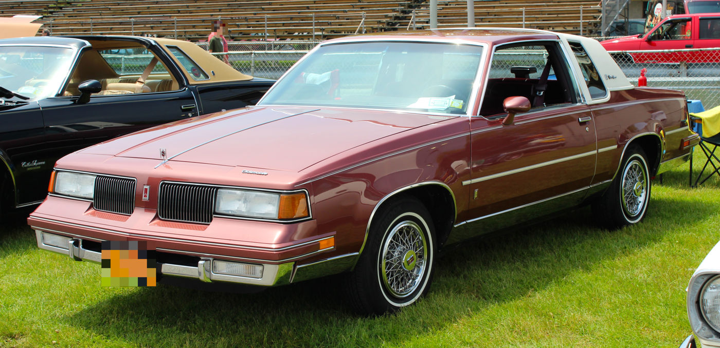 Front three-quarter view of a rose-coloured 1988 Oldsmobile Cutlass Supreme coupe with a white vinyl roof, vertical-slat grilles, whitewall tyres and wire wheel covers, parked on grass