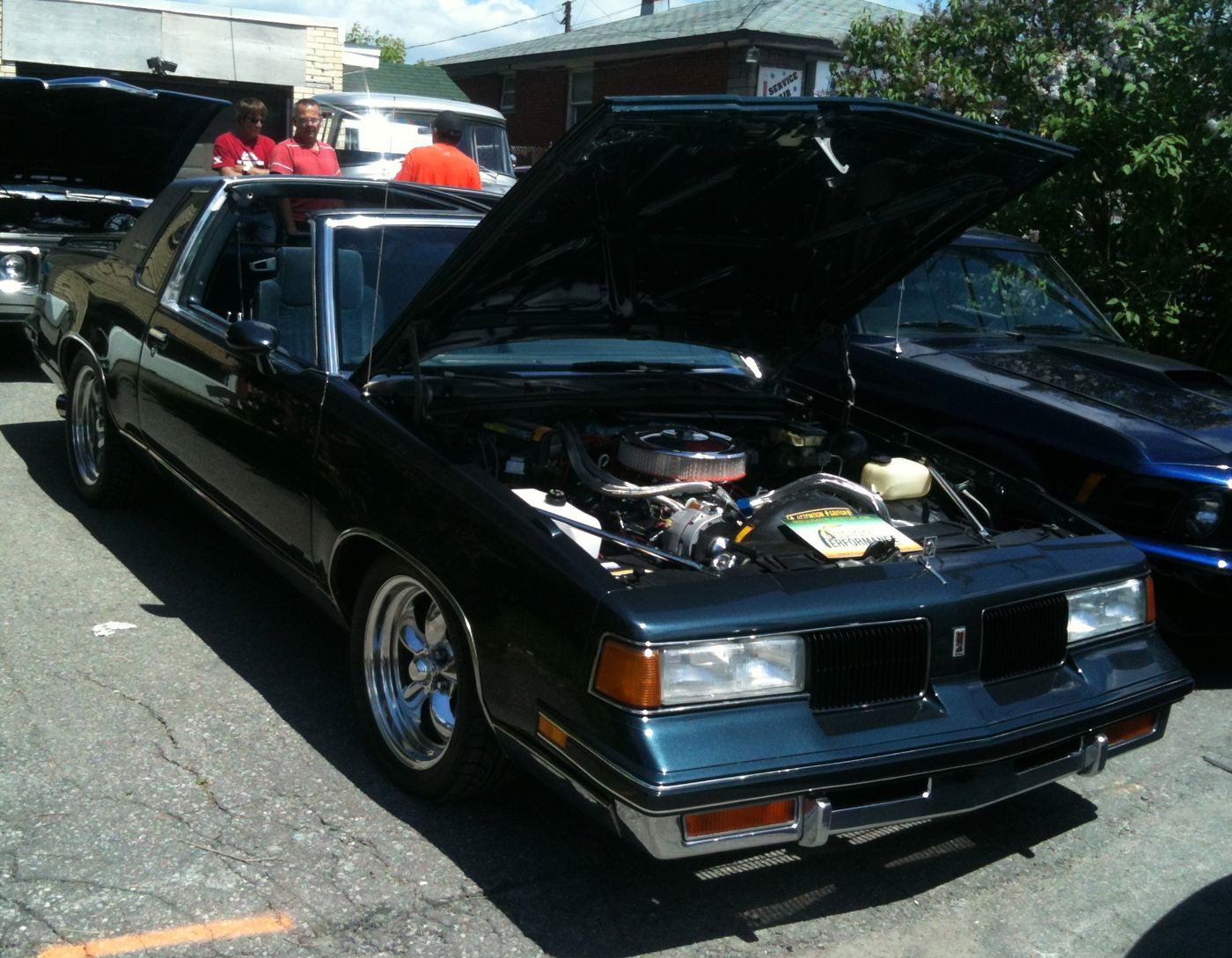 Front three-quarter view of a dark teal 1988 Oldsmobile Cutlass Supreme Classic coupe with its hood raised on a modified engine and chrome wheels