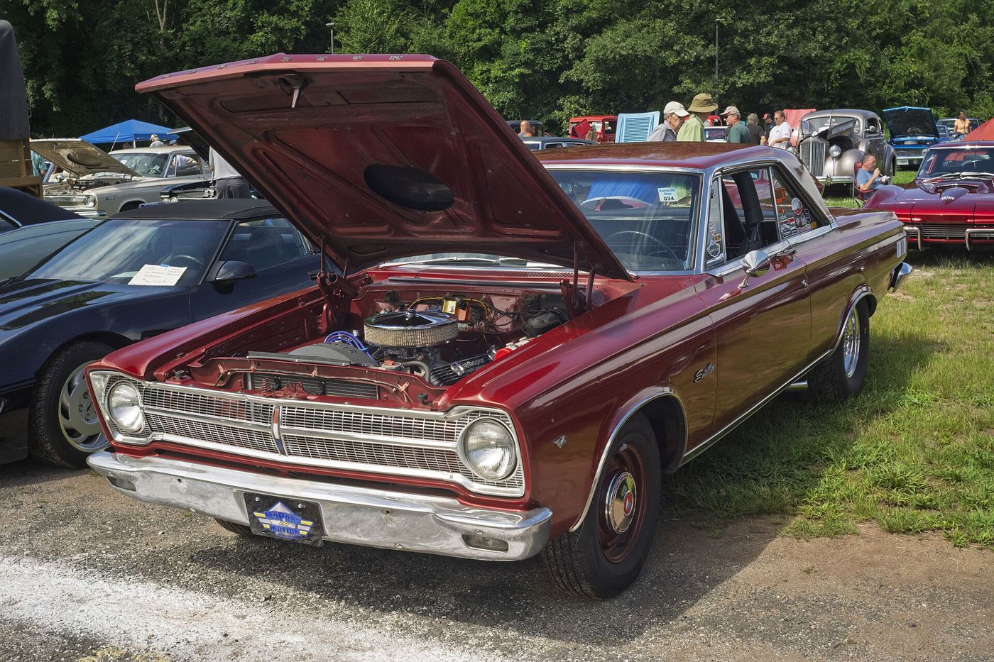 Red 1965 Plymouth Satellite hardtop at a car show with the hood raised over its V8, showing the 1965 grille and single headlamps