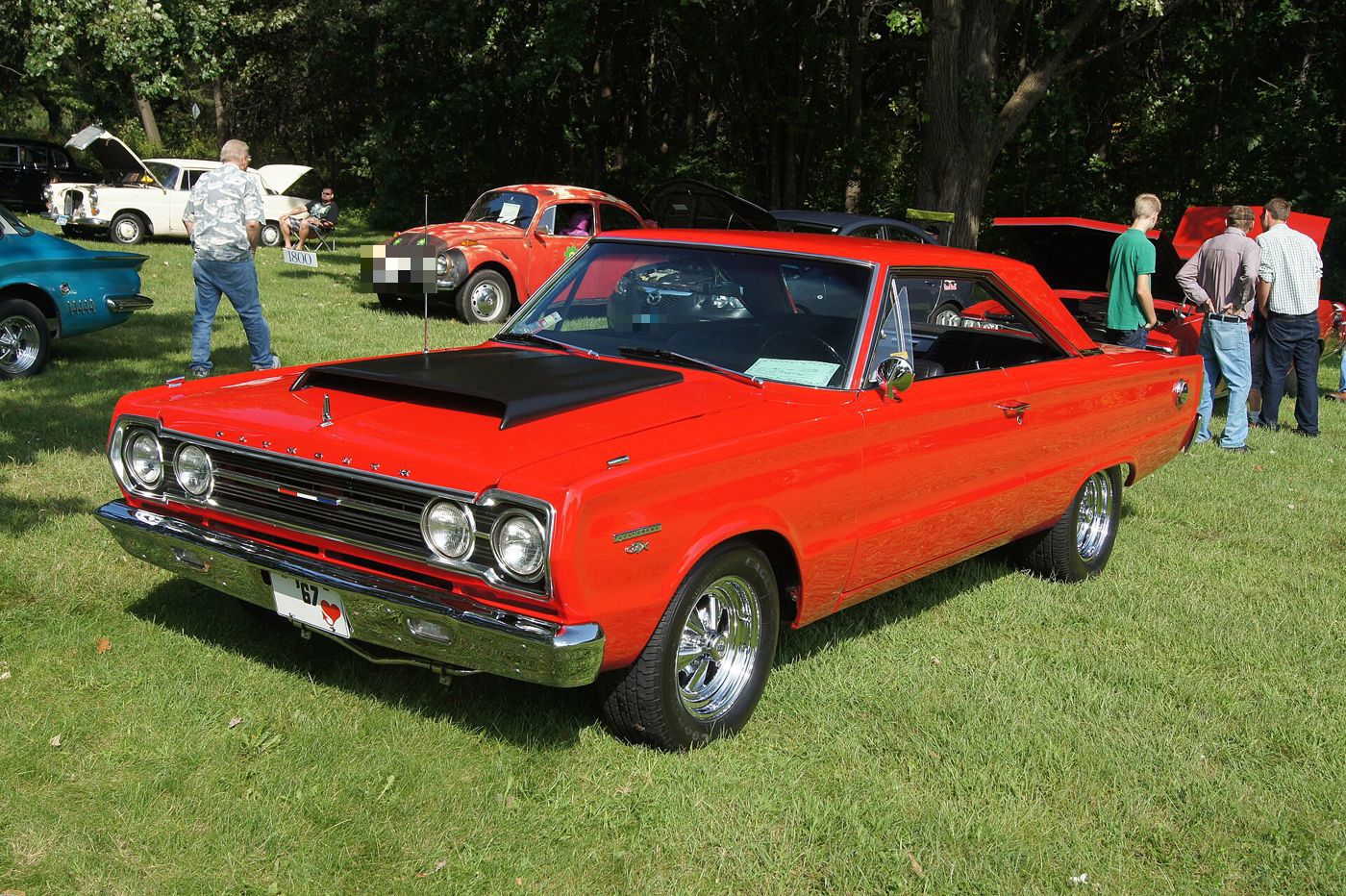 Red 1967 Plymouth Belvedere GTX with black hood and quad headlamps, front three-quarter on grass at a car show