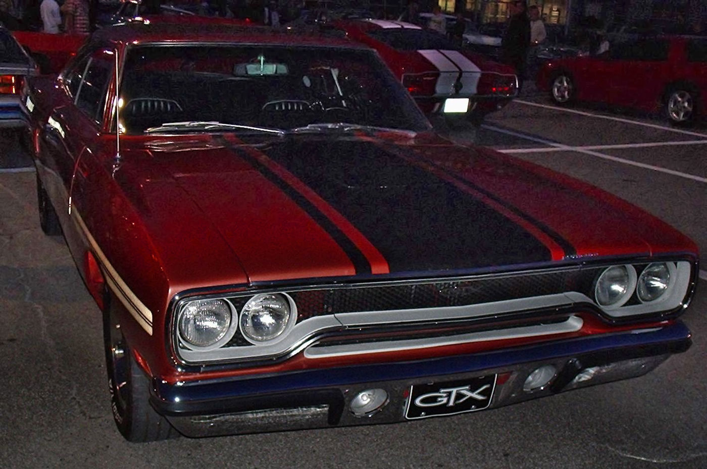 Red 1970 Plymouth GTX seen close from the front at night, showing the 1970 loop grille and black-striped hood