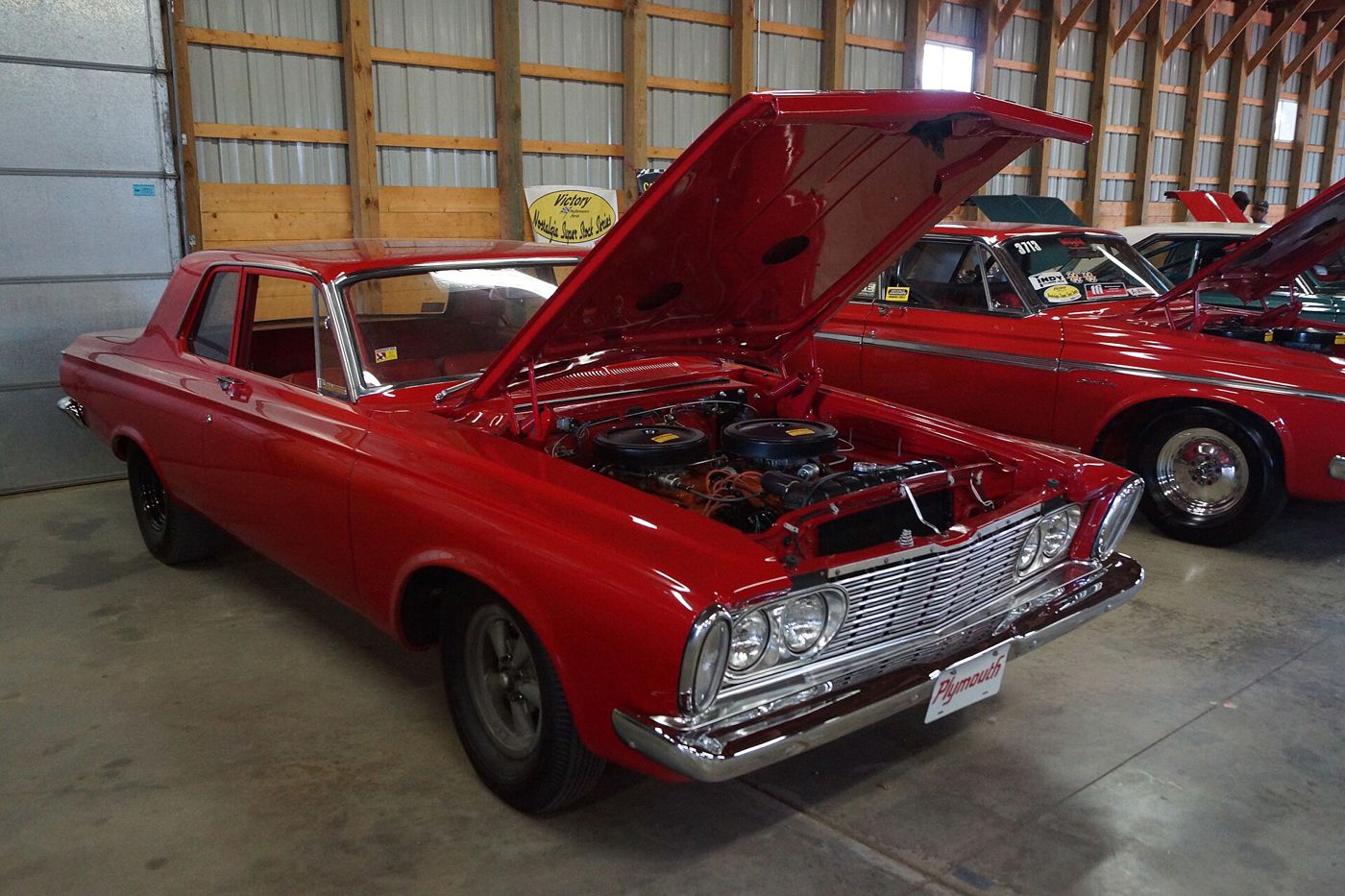 Red 1963 Plymouth Savoy two-door sedan with its hood raised over the Max Wedge V8, in an indoor show barn