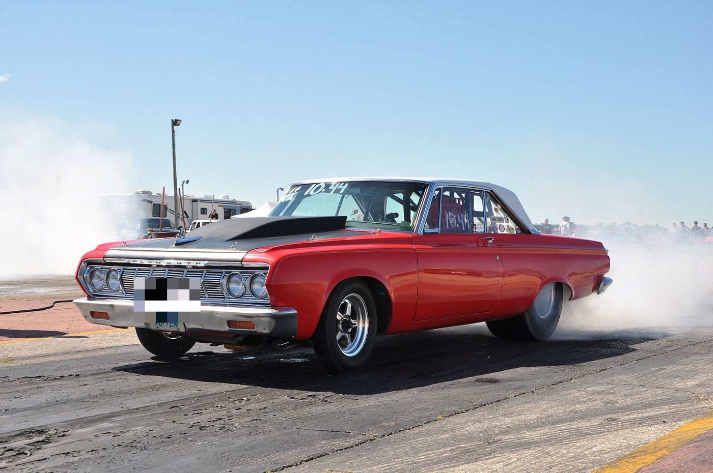 Red 1964 Plymouth two-door sedan with a black hood scoop spinning its rear tyres in a cloud of smoke on a drag strip