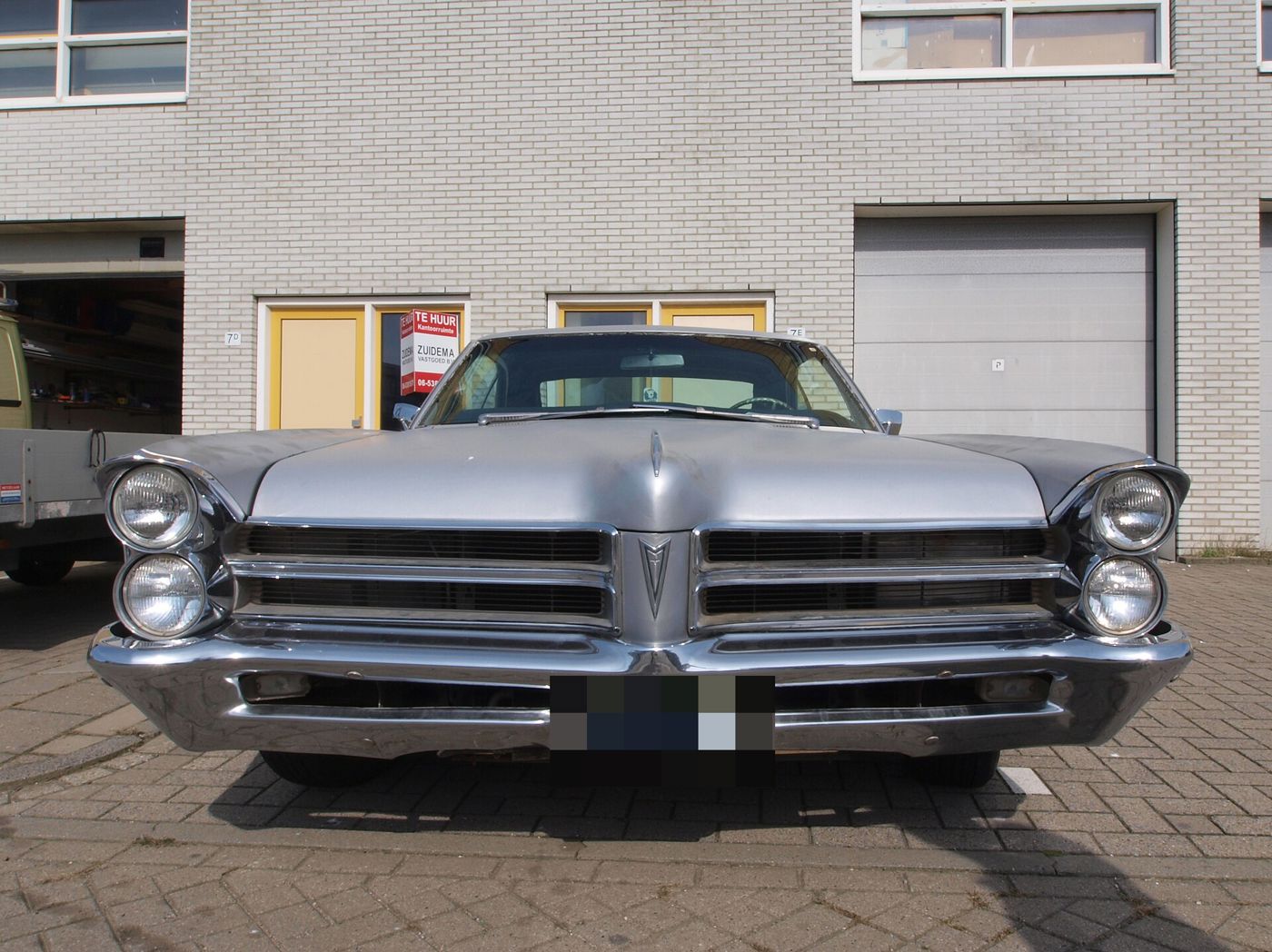 Silver 1965 Catalina seen head-on, showing its stacked headlights and split grille, parked in front of a brick building