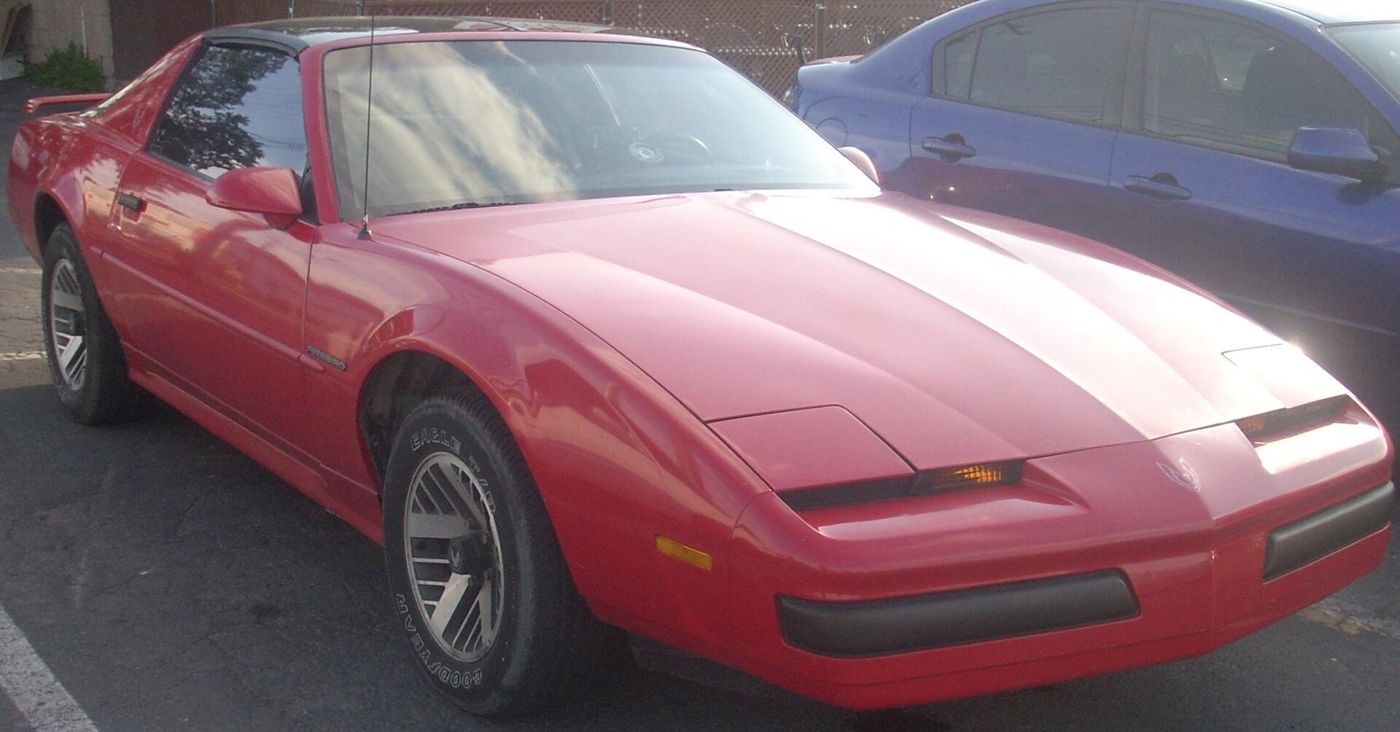 Red third-generation Pontiac Firebird with hidden headlamps and turbo-style wheels, front three-quarter in a parking lot