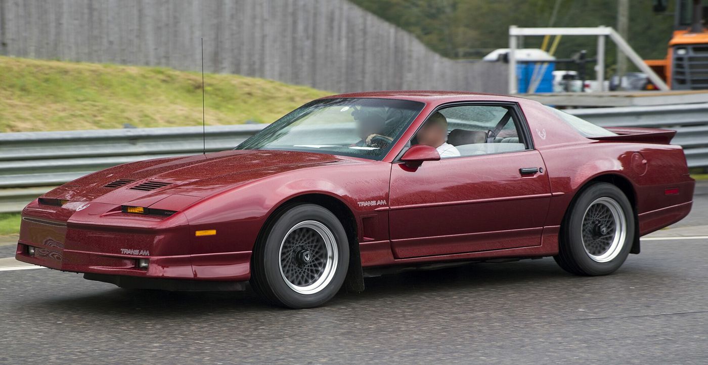 Dark red 1987 Trans Am with ground-effects skirts and lattice wheels in side view on a racetrack paddock road