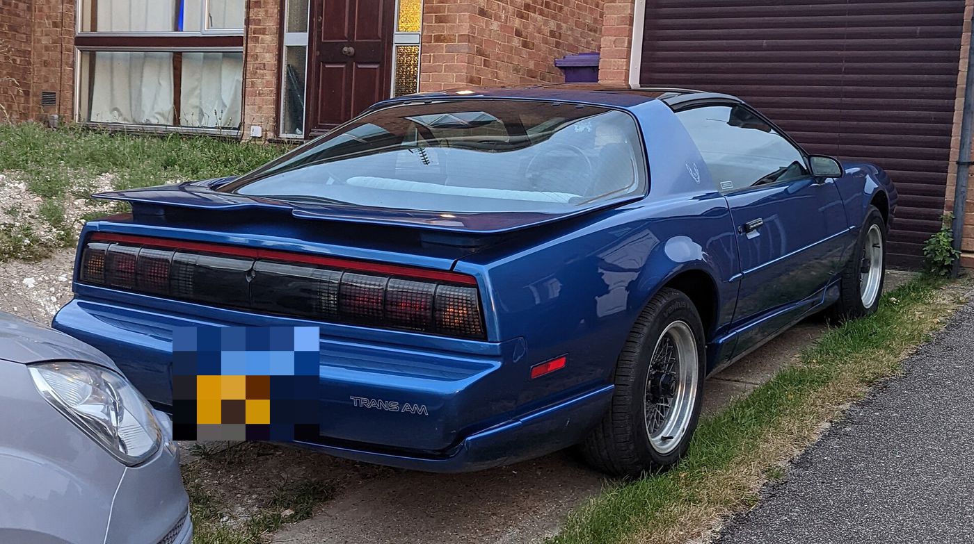 Blue Trans Am from the rear three-quarter showing its full-width taillamp panel, rear spoiler and TRANS AM lettering, parked by a house in England