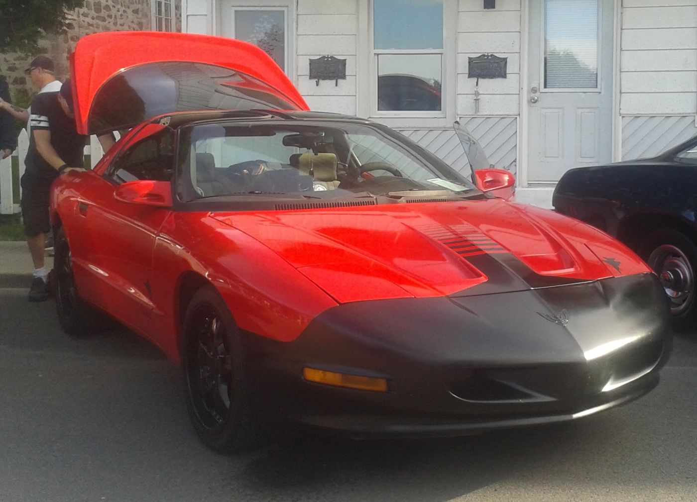 Red 1995 Firebird with a dark nose stripe and T-tops, front three-quarter at a car show, a spectator at the open hatch of the car behind