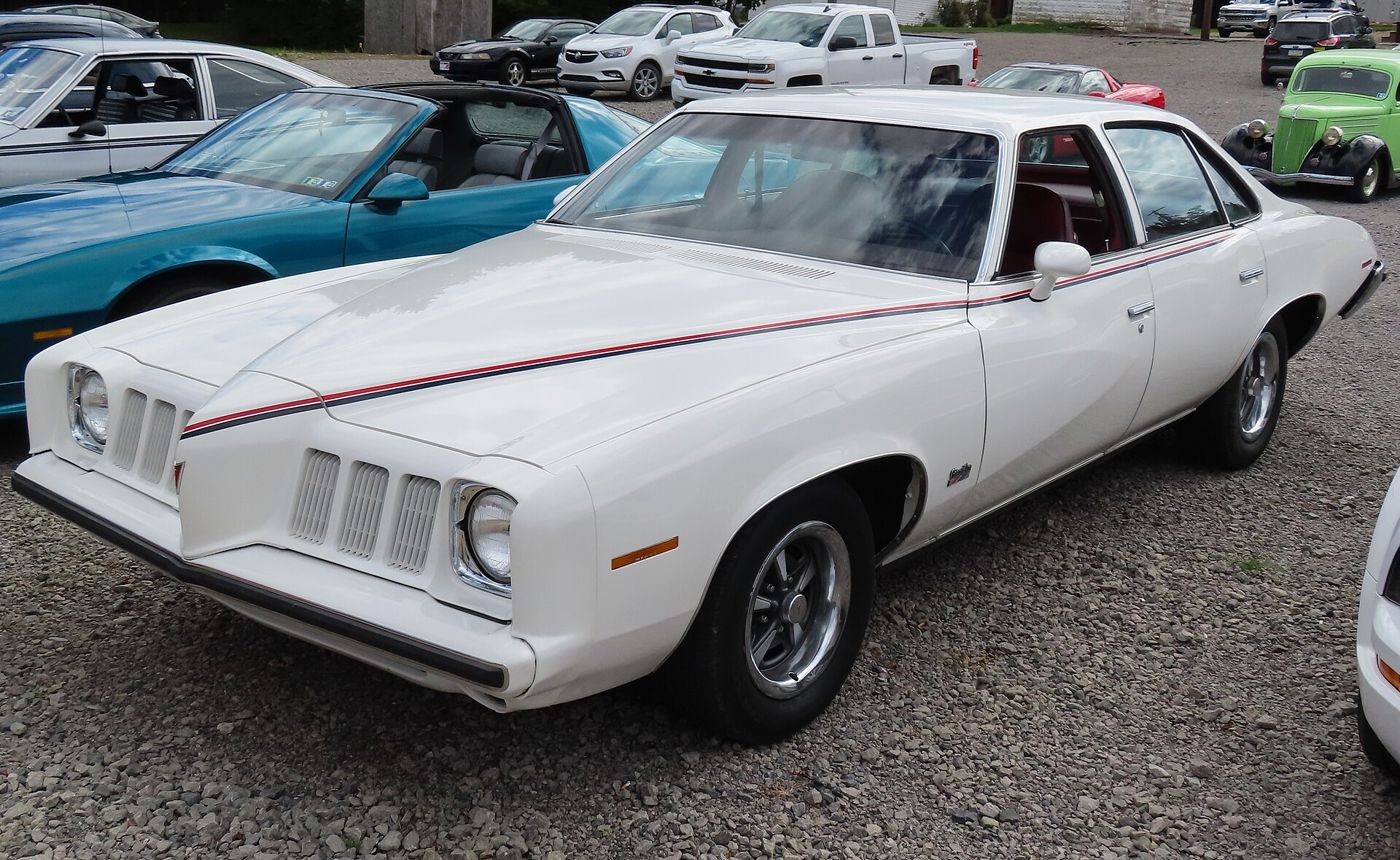 White 1973 Pontiac Grand Am four-door Colonnade hardtop with its flexible Endura nose, split vertical-bar grilles, red and blue stripe and Rally II wheels, front three-quarter on gravel