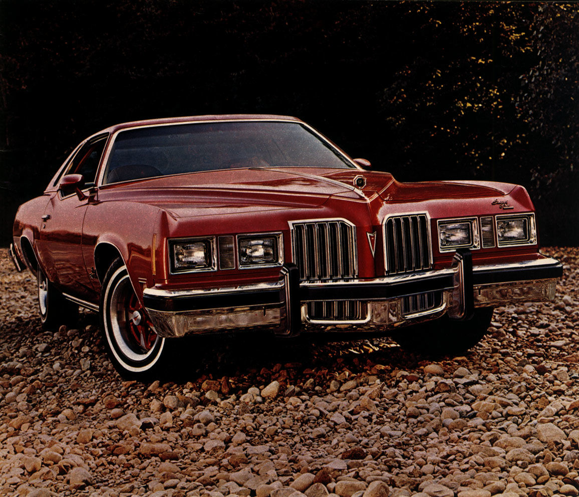 Red 1977 Grand Prix with a white landau roof and tall chrome grille, front three-quarter on a gravel bed