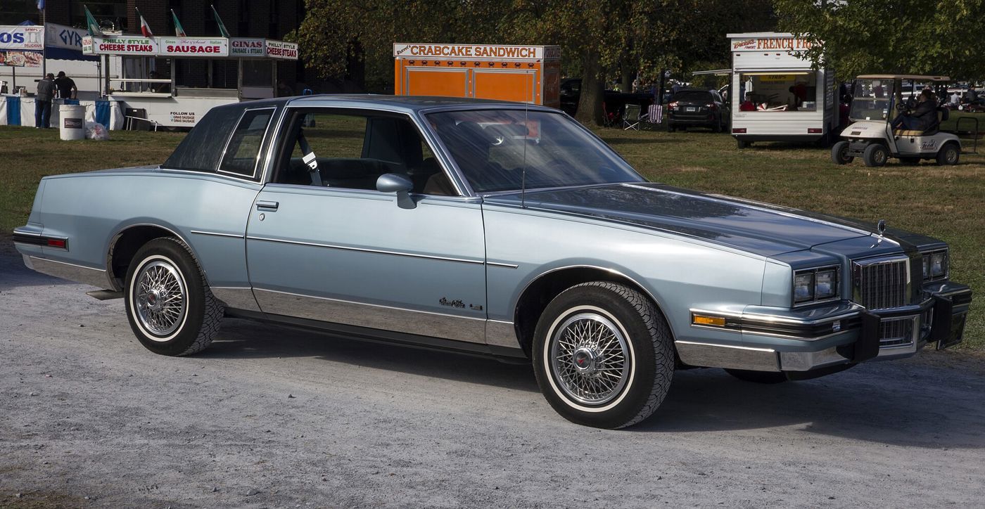 Two-tone light and dark blue 1985 Grand Prix LE coupe with wire wheel covers, front three-quarter on gravel at a show
