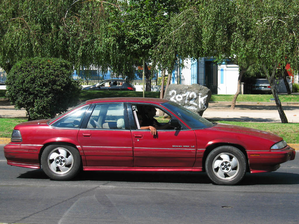 Red 1992 Grand Prix LE four-door sedan driving past trees, in side view