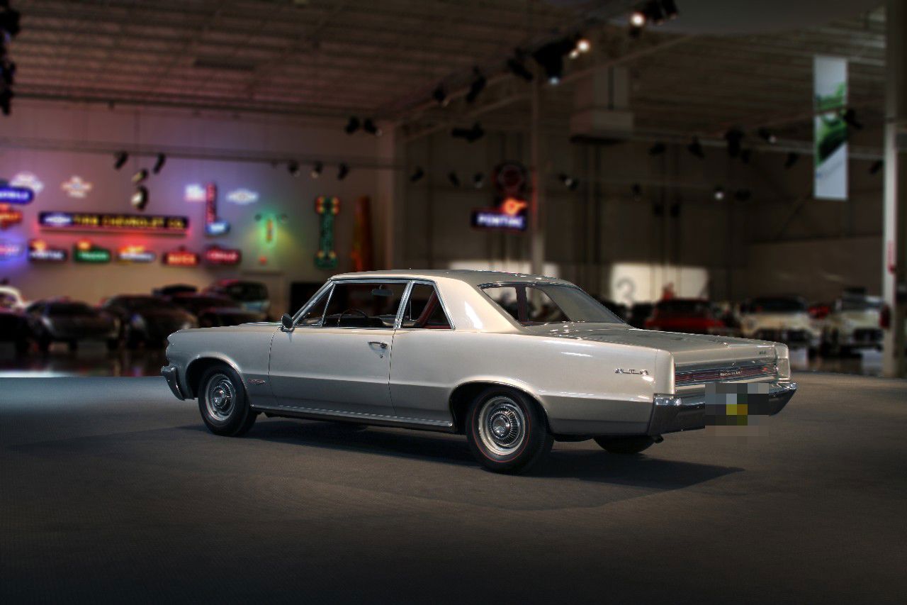 Silver 1964 GTO coupe seen from the rear three-quarter in the GM Heritage Center, neon signs and collection cars behind