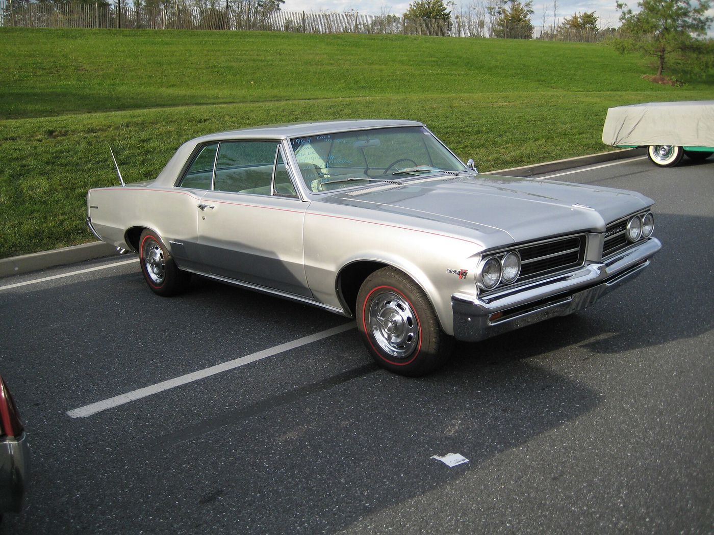 Silver 1964 Pontiac Tempest LeMans hardtop with red pinstripe, stacked quad headlamps and chrome wheels with redline tires, front three-quarter in a parking lot