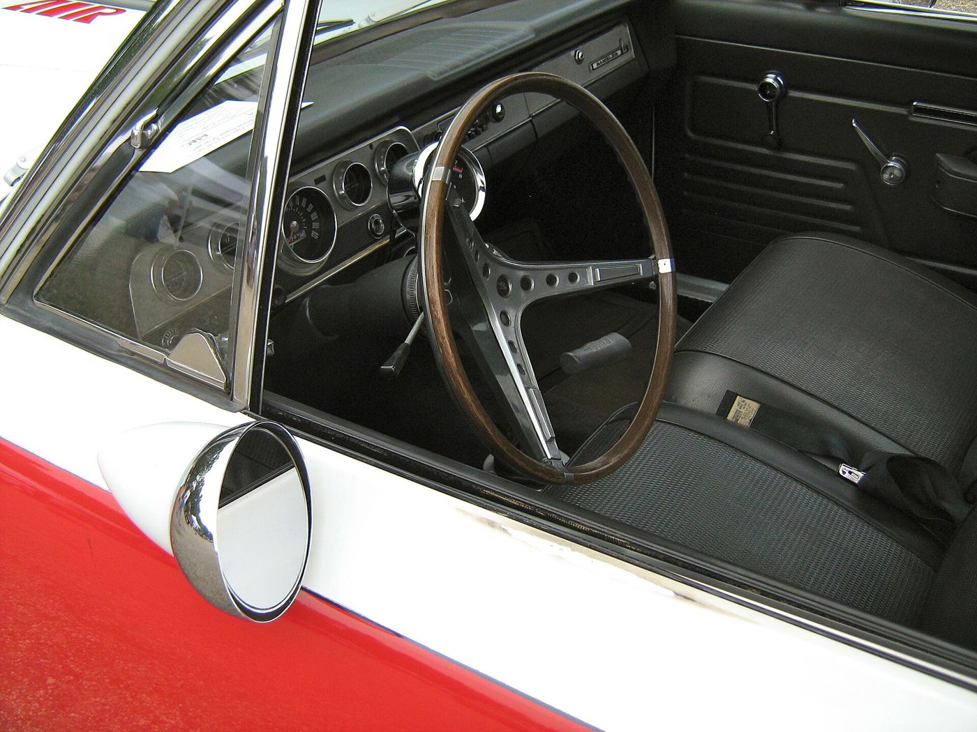 View through the open door of an SC/Rambler: woodgrain steering wheel, dash and black vinyl seat, with the chrome racing mirror in the foreground