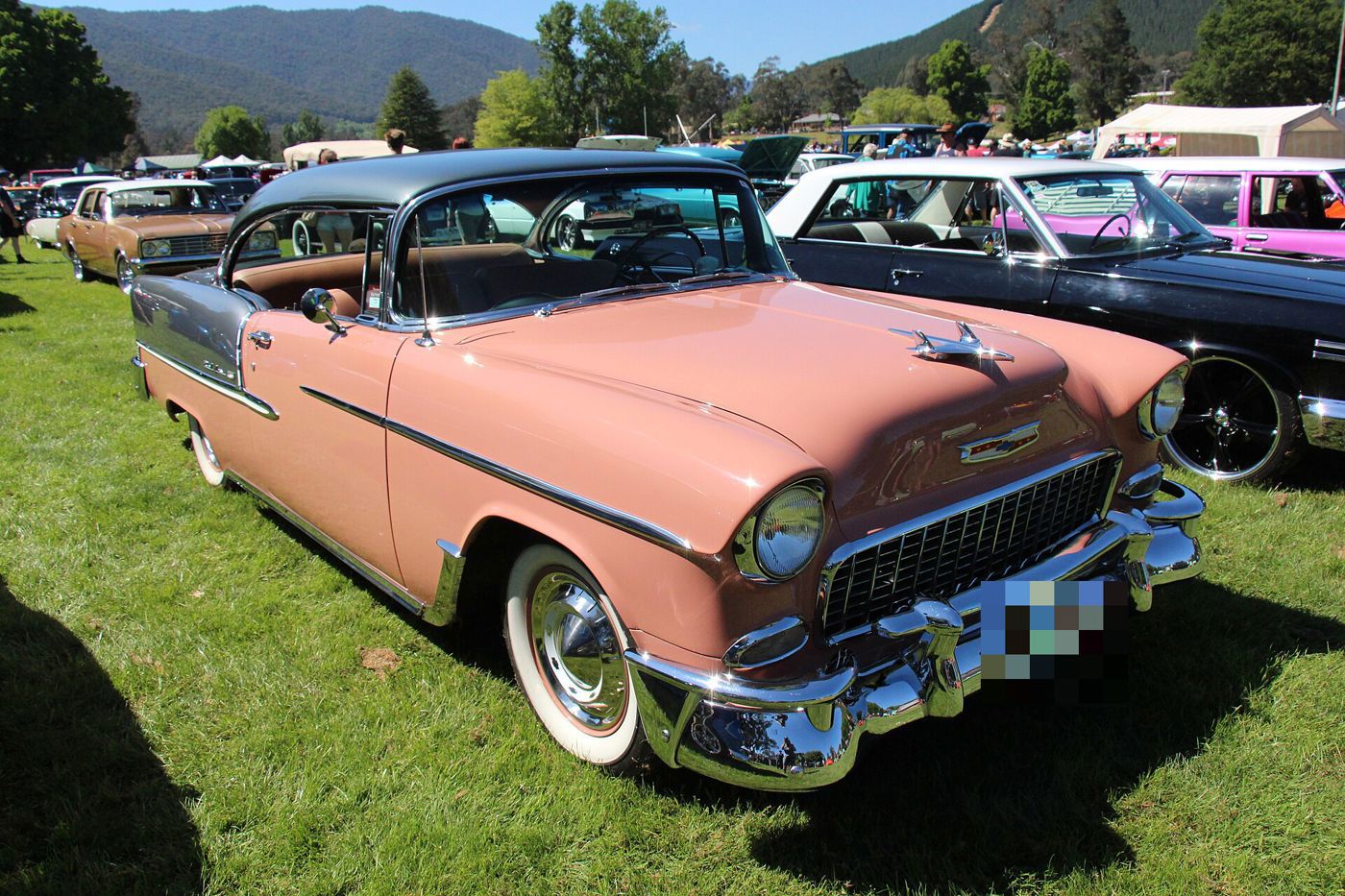 Coral 1955 Chevrolet Bel Air hardtop with a dark roof, seen from the front three-quarter on grass at a car show