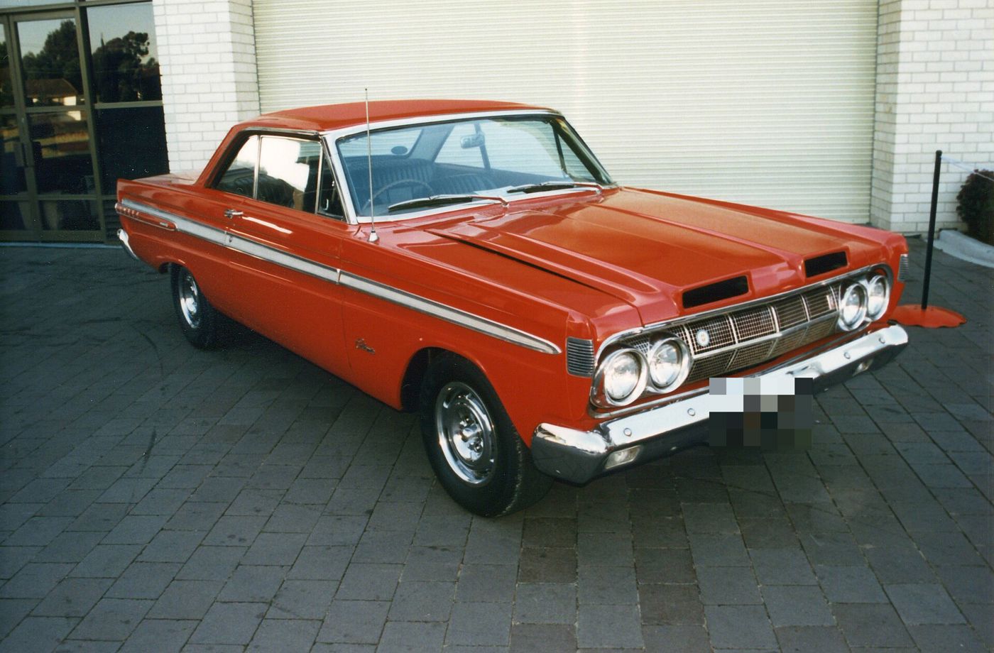 Red 1964 Mercury Comet Cyclone hardtop with chrome wheel covers, front three-quarter in front of a building (scanned print)