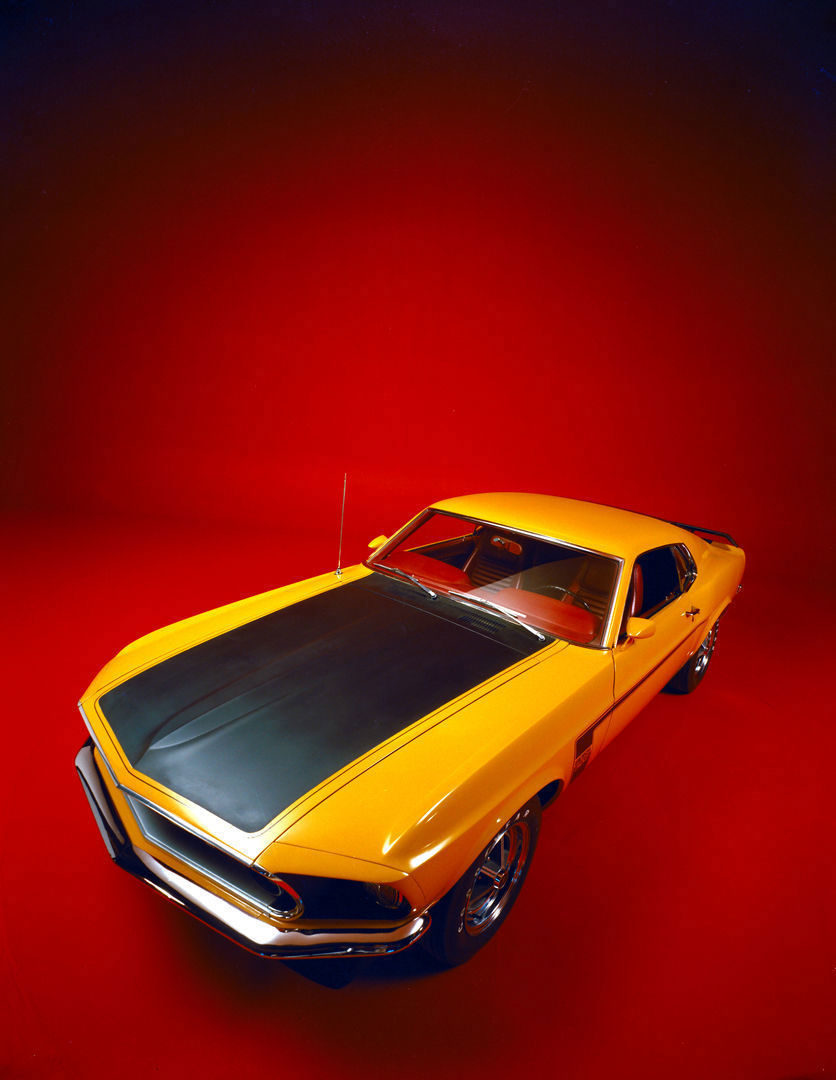 High view of a yellow 1969 Mustang Boss 302 with a matte black hood on a red studio backdrop