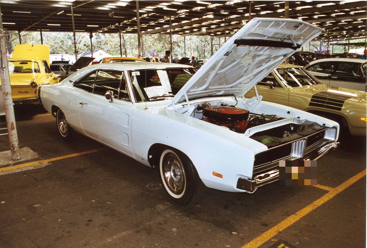 White 1969 Dodge Charger R/T with its hood open over an orange-painted engine, front three-quarter under a shade roof (scanned print)
