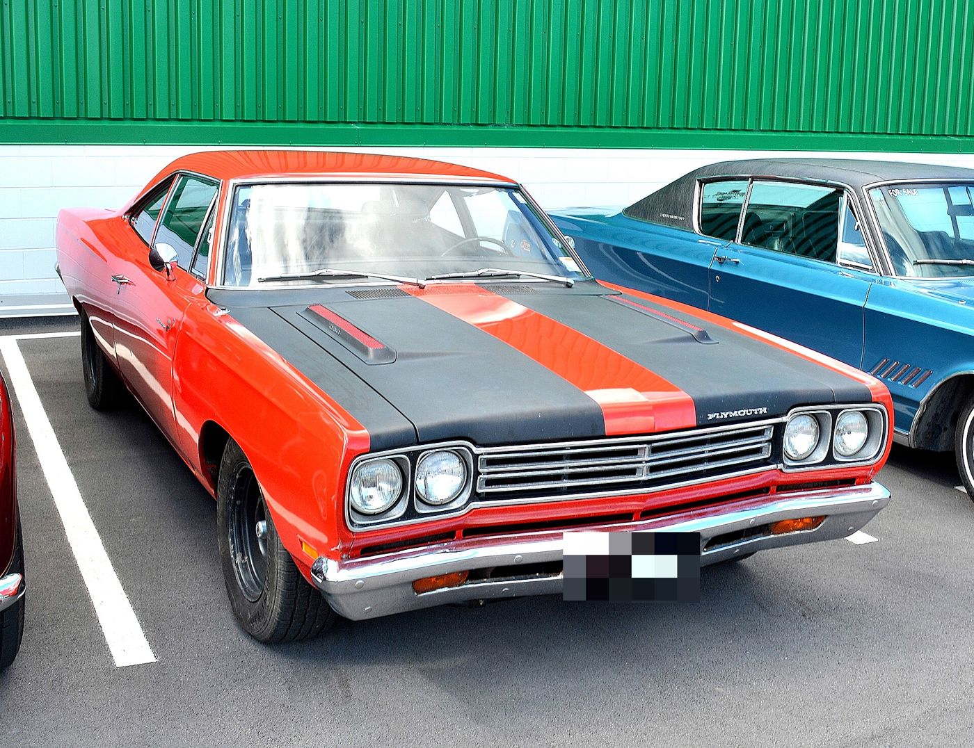 Red-orange 1969 Plymouth Road Runner with a black hood and hood stripe and black steel wheels, front three-quarter in a parking lot