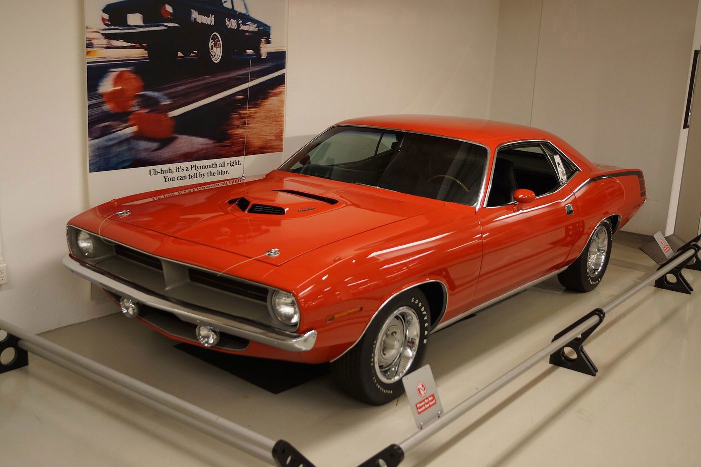 Red-orange 1970 Plymouth Hemi Cuda hardtop with shaker hood scoop on display in front of a period Plymouth advertisement