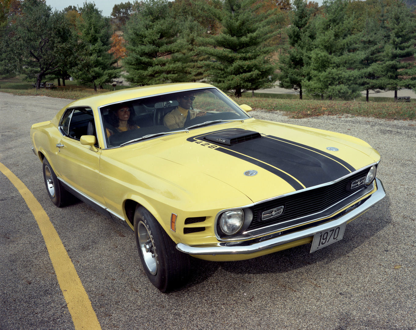 Yellow 1970 Ford Mustang Mach 1 with black hood stripe and shaker scoop, parked in a lot with pine trees and a 1970 display plate