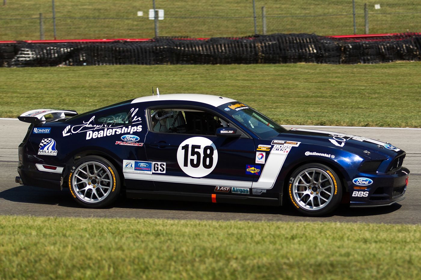 Dark blue and white Mustang Boss 302R race car number 158 with a rear wing and sponsor decals, seen side-on on the Mid-Ohio circuit