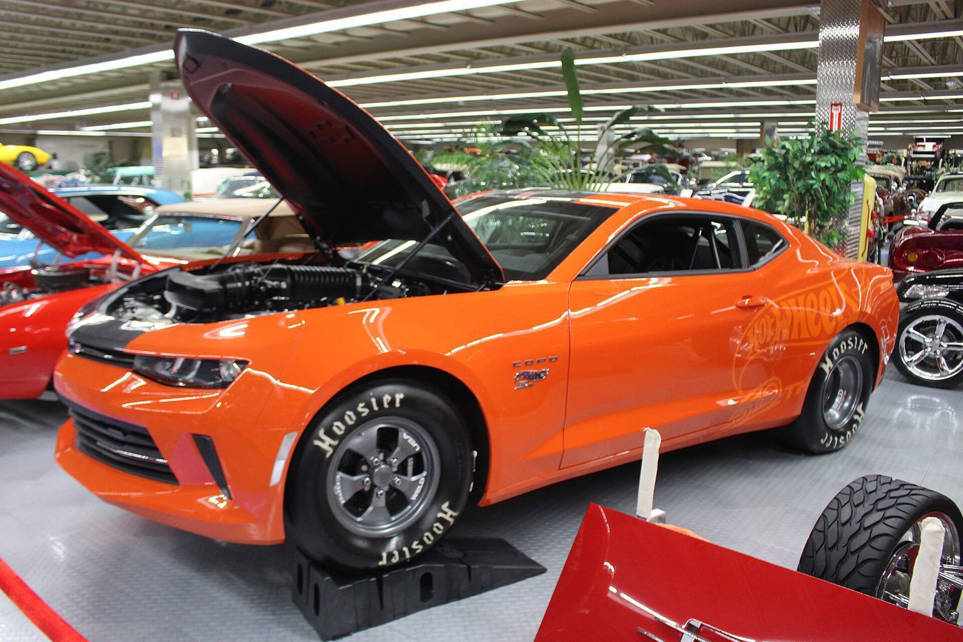 Orange COPO Camaro drag car with its hood up over a supercharged engine and Hot Wheels graphics, on display
