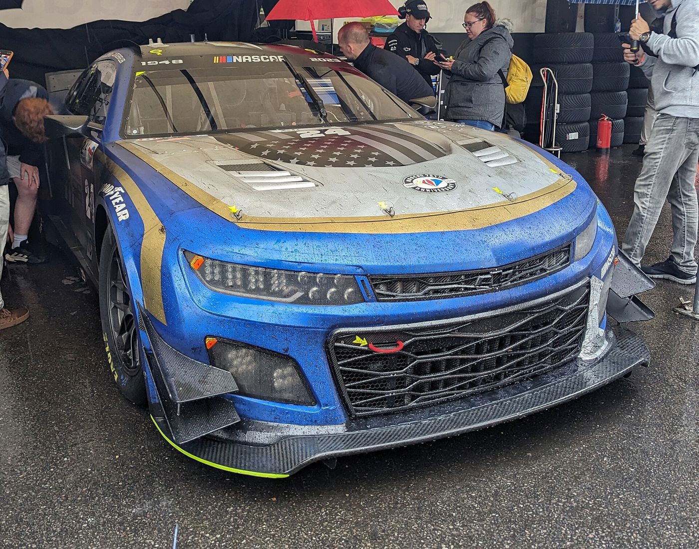 Blue, white and gold number 24 Garage 56 Camaro with rain on its hood, in a paddock tent