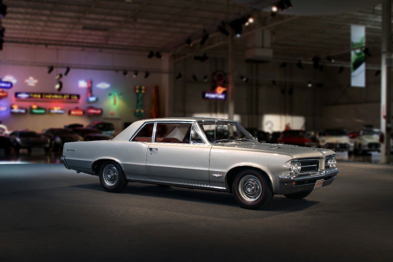 Silver 1964 Pontiac GTO hardtop shown from the side in the dimly lit GM Heritage Center hall, neon signs behind