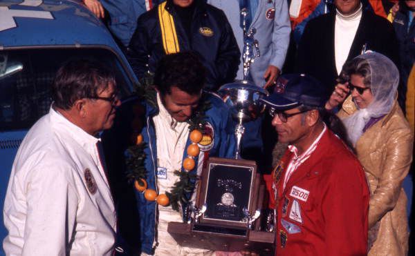 Richard Petty wearing an orange lei holds a trophy while Lt. Gov. Tom Adams in a red jacket presents it