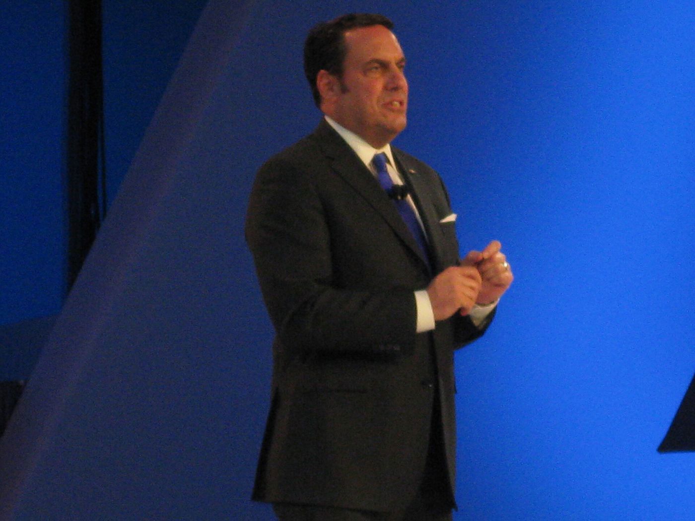 Mark Reuss in a dark suit and blue tie speaking on a stage lit blue