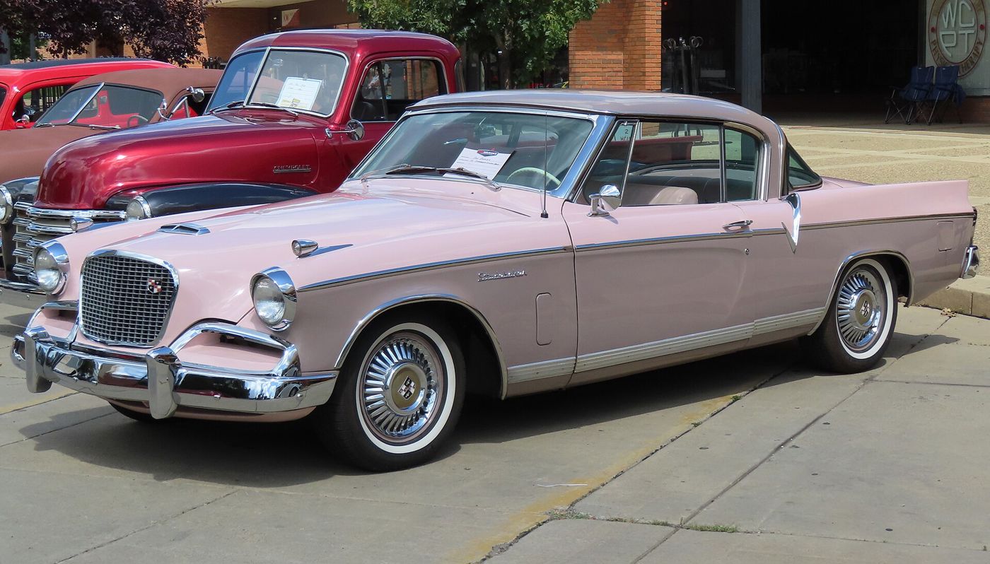 Pink two-tone 1956 Studebaker Golden Hawk hardtop with its tall mesh grille, hood scoop, whitewall tires and tailfins, front three-quarter at a car show
